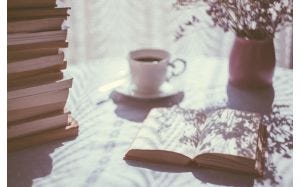 A table with a stack of books, an open books, a pot of flowers, and a mug of tea