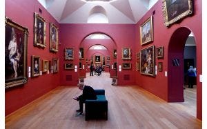 Interior of an art gallery, with red walls and gold framed paintings